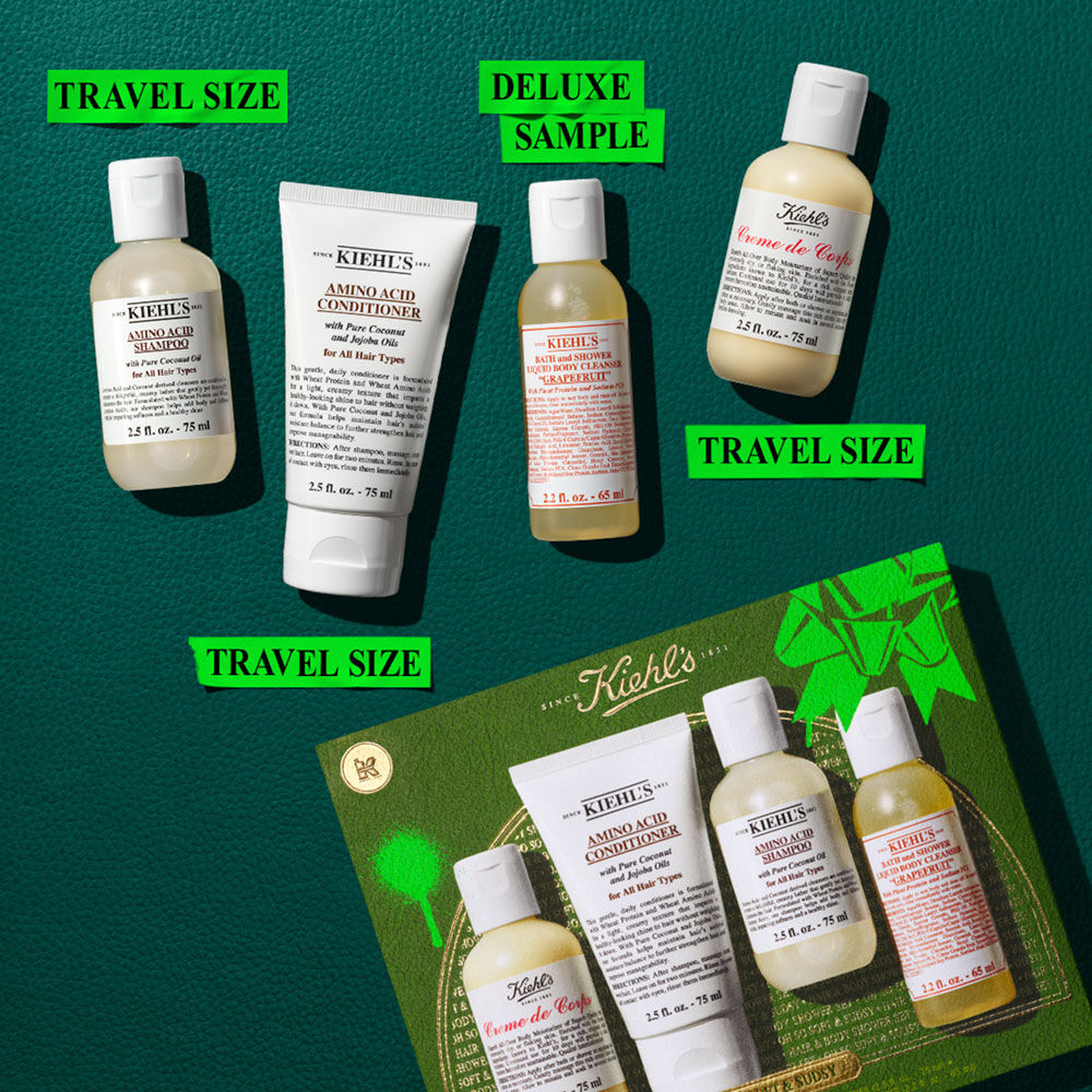 Una imagen plana sobre fondo verde oscuro muestra un set Kiehl's con precio. Incluye cuatro productos de viaje: champu, acondicionador, limpiador corporal (pomelo) y Crema de Corps. Una caja de regalo verde es visible abajo a la derecha.