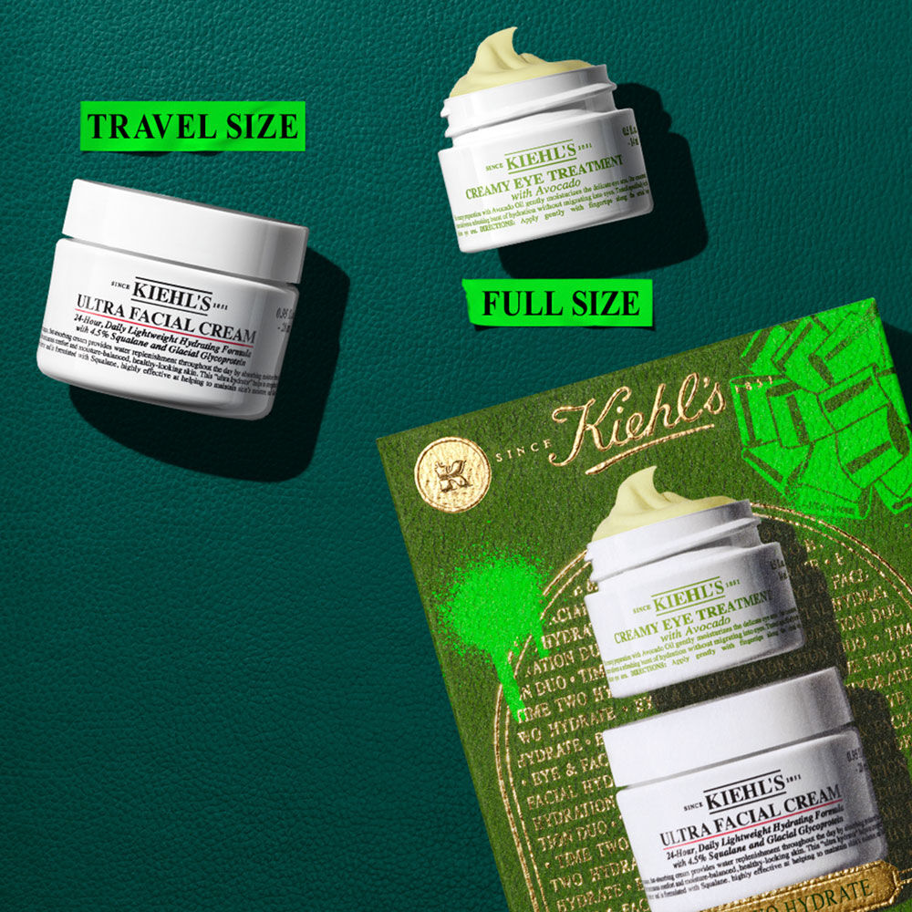 Un grafico muestra un tarro blanco de "KIEHL’S ULTRA FACIAL CREAM" etiquetado como "TAMANO DE VIAJE", y un tarro abierto de "KIEHL’S CREAMY EYE TREATMENT WITH AVOCADO" etiquetado como "TAMANO COMPLETO". Ambos sobre fondo verde texturizado. Una caja de regalo Kiehl’s verde y dorada parcial abajo a la derecha.