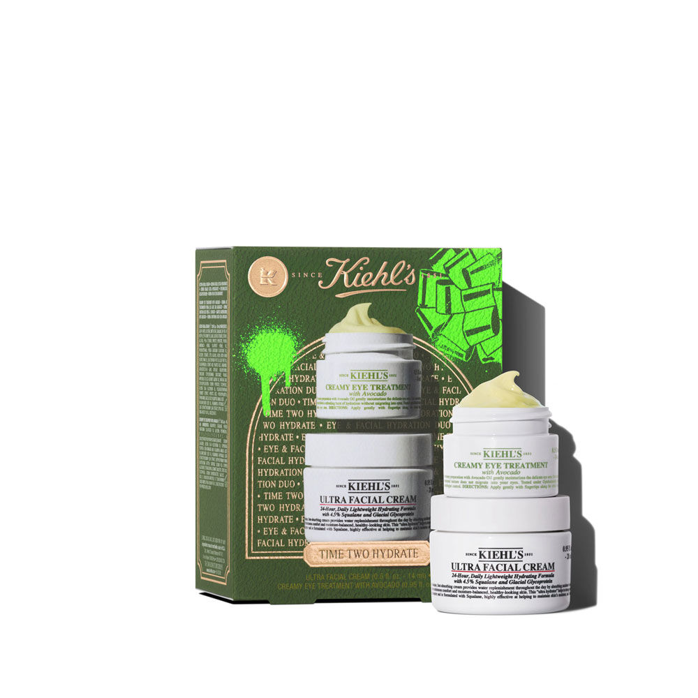 Una caja de regalo Kiehl's verde a la izquierda, con texto "TIME TWO HYDRATE" y elementos graficos verdes. A la derecha, dos tarros apilados: el superior es "KIEHL'S CREAMY EYE TREATMENT with avocado" con remolino amarillo-verde visible. El inferior es "KIEHL'S ULTRA FACIAL CREAM".