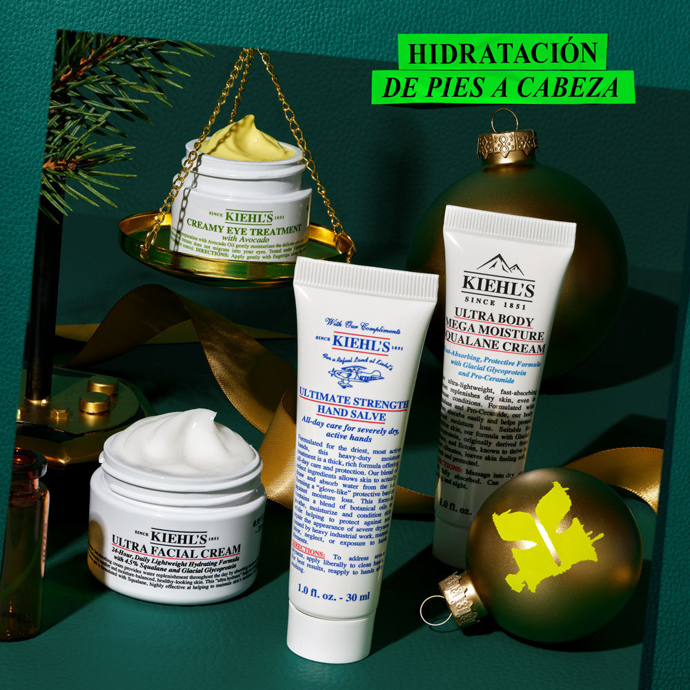 Grafico festivo con fondo verde oscuro texturizado. Muestra varios productos Kiehl's y decoracion navidena. Un tarro abierto de "CREAMY EYE TREATMENT WITH AVOCADO" en una balanza dorada. Tambien "ULTRA FACIAL CREAM", "ULTIMATE STRENGTH HAND SALVE" y "ULTRA BODY MEGA MOISTURE SQUALANE CREAM". Un adorno dorado con logo "K". Una caja verde dice: "Give the gift of HEAD-TO-TOE HYDRATION this holiday!".