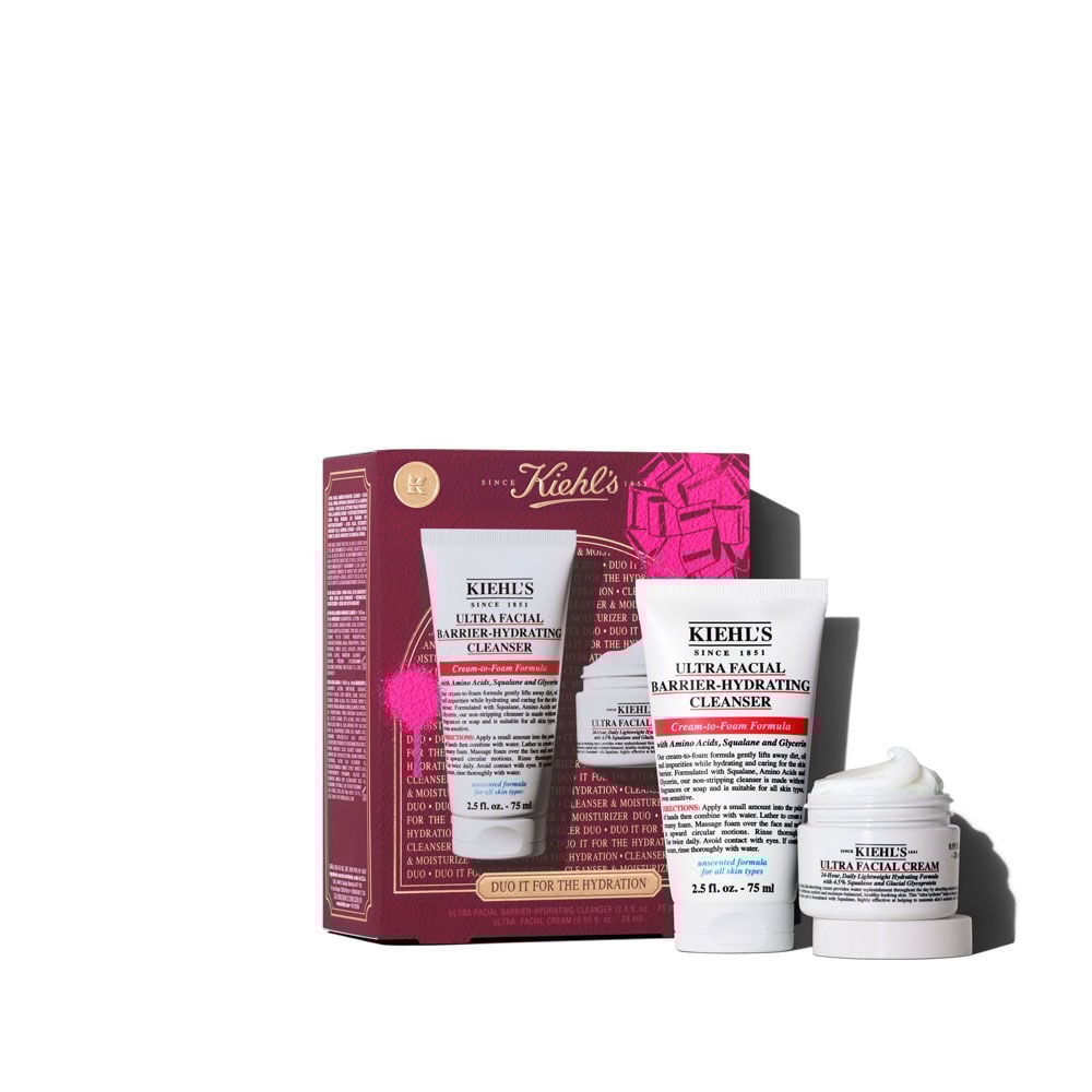 Un grafico muestra una caja de regalo Kiehl's, mayormente roja oscura, con un lazo rosa arriba a la derecha y una salpicadura rosa abajo a la izquierda. La caja dice "DUO IT FOR THE HYDRATION" y "ULTRA FACIAL BARRIER-HYDRATING CLENSER + ULTRA FACIAL CREAM". A la derecha, se ven los dos productos fuera de su empaque.
