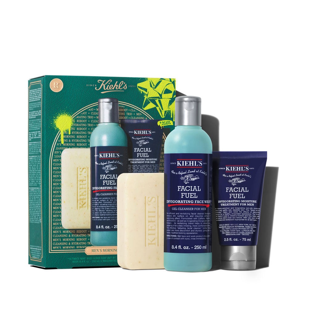Un grafico muestra una caja Kiehl’s verde oscuro con detalles blancos y dorados. Contiene tres productos: jabon corporal, gel limpiador y humectante facial. La caja, con salpicaduras y lazo verde, dice "MEN’S MORNING REBOOT".