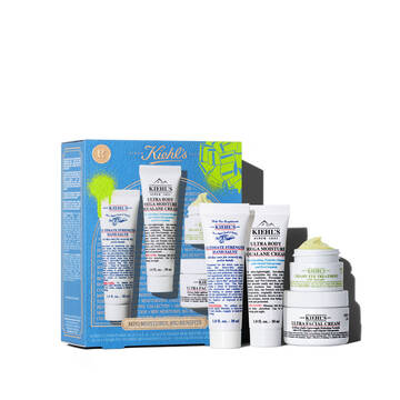 Un grafico muestra una caja de regalo Kiehl's azul con detalles verdes a la izquierda. Contiene cuatro productos: "KIEHL’S ULTIMATE STRENGTH HAND SALVE", "KIEHL’S ULTRA BODY MEGA MOISTURE SQUALANE CREAM", "KIEHL’S CREAMY EYE TREATMENT WITH AVOCADO" y "KIEHL’S ULTRA FACIAL CREAM". La caja tiene salpicaduras verdes y un lazo, con el texto "MINI HIDRATACION, GRANDES BENEFICIOS".