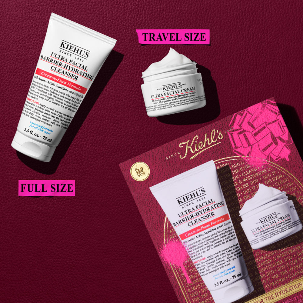 Un grafico muestra un tubo blanco de "KIEHL'S ULTRA FACIAL BARRIER-HYDRATING CLEANSER" (TAMANO COMPLETO) y un tarro blanco mas pequeno de "KIEHL'S ULTRA FACIAL CREAM" (TAMANO DE VIAJE). Arriba a la derecha, una caja rosa salpicada. Una caja de regalo Kiehl's parcial con un lazo rosa visible abajo a la derecha.