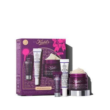 Un grafico con una caja de regalo Kiehl’s morada y detalles dorados. Contiene tres productos: "Kiehl's Super Multi-Corrective Eye Zone Treatment", "Kiehl's Retinol Skin-Renewing Daily Micro-Dose Serum" y "Kiehl's Super Multi-Corrective Cream" visible. La caja tiene salpicaduras rosas y un mono, con el texto "LET IT SMOOOOOTH".