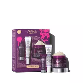 Un grafico con una caja de regalo Kiehl’s morada y detalles dorados. Contiene tres productos: "Kiehl's Super Multi-Corrective Eye Zone Treatment", "Kiehl's Retinol Skin-Renewing Daily Micro-Dose Serum" y "Kiehl's Super Multi-Corrective Cream" visible. La caja tiene salpicaduras rosas y un mono, con el texto "LET IT SMOOOOOTH".