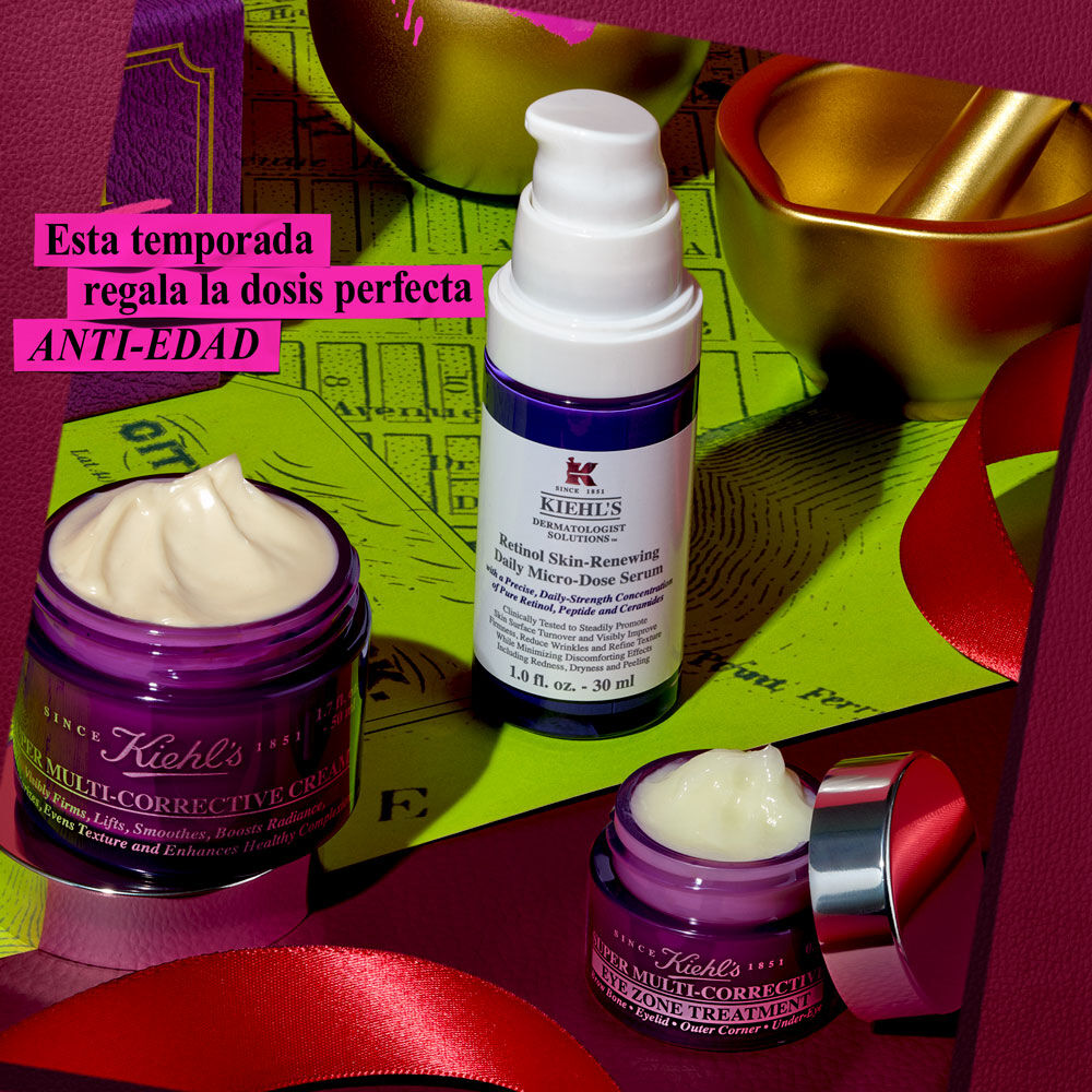 Un grafico festivo presenta tres productos antienvejecimiento de Kiehl's: un tarro morado de "Kiehl's Super Multi-Corrective Cream", una botella blanca y morada de "Kiehl's Retinol Skin-Renewing Daily Micro-Dose Serum" y un tarro morado mas pequeno de "Kiehl's Super Multi-Corrective Eye Zone Treatment". Estan dispuestos sobre una superficie verde, con cintas rojas y un mortero y una mano de oro en el fondo. Un cuadro de texto rosa en la esquina superior izquierda dice: "¡Regala ANTI-EDAD esta temporada!".