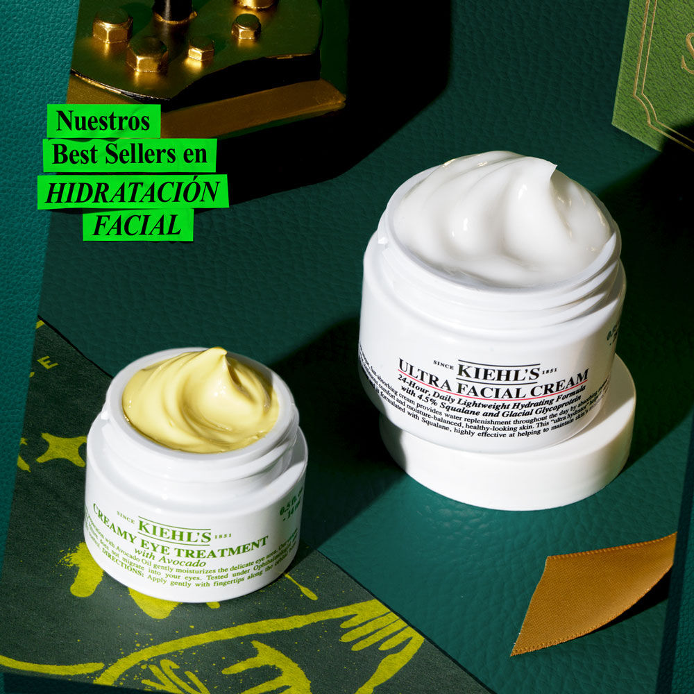 Un grafico muestra un tarro blanco de "KIEHL'S CREAMY EYE TREATMENT with avocado" con un remolino amarillo-verde, y un tarro blanco grande de "KIEHL'S ULTRA FACIAL CREAM" con remolino blanco. Sobre superficie verde oscuro con acentos dorados y un objeto decorativo. Texto verde: "¡Regala HIDRATACION FACIAL esta Navidad!".