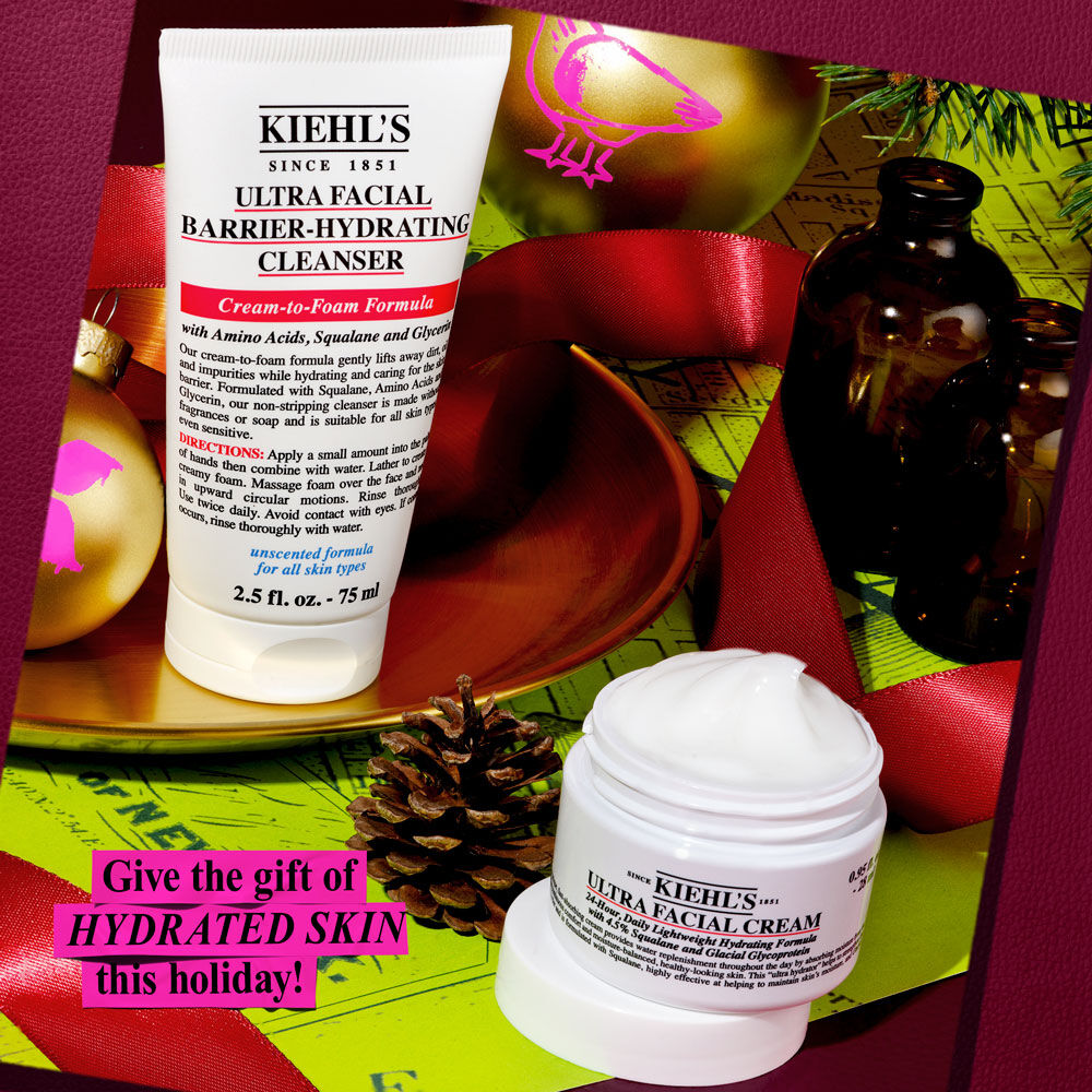 AUn grafico festivo muestra un tubo blanco de "Kiehl’s Ultra Facial Barrier-Hydrating Cleanser" y un tarro blanco de "Kiehl’s Ultra Facial Cream". Fondo con cinta roja y papel de regalo verde con adornos dorados. Un texto prominente dice: "¡Regala PIEL HIDRATADA esta Navidad!".