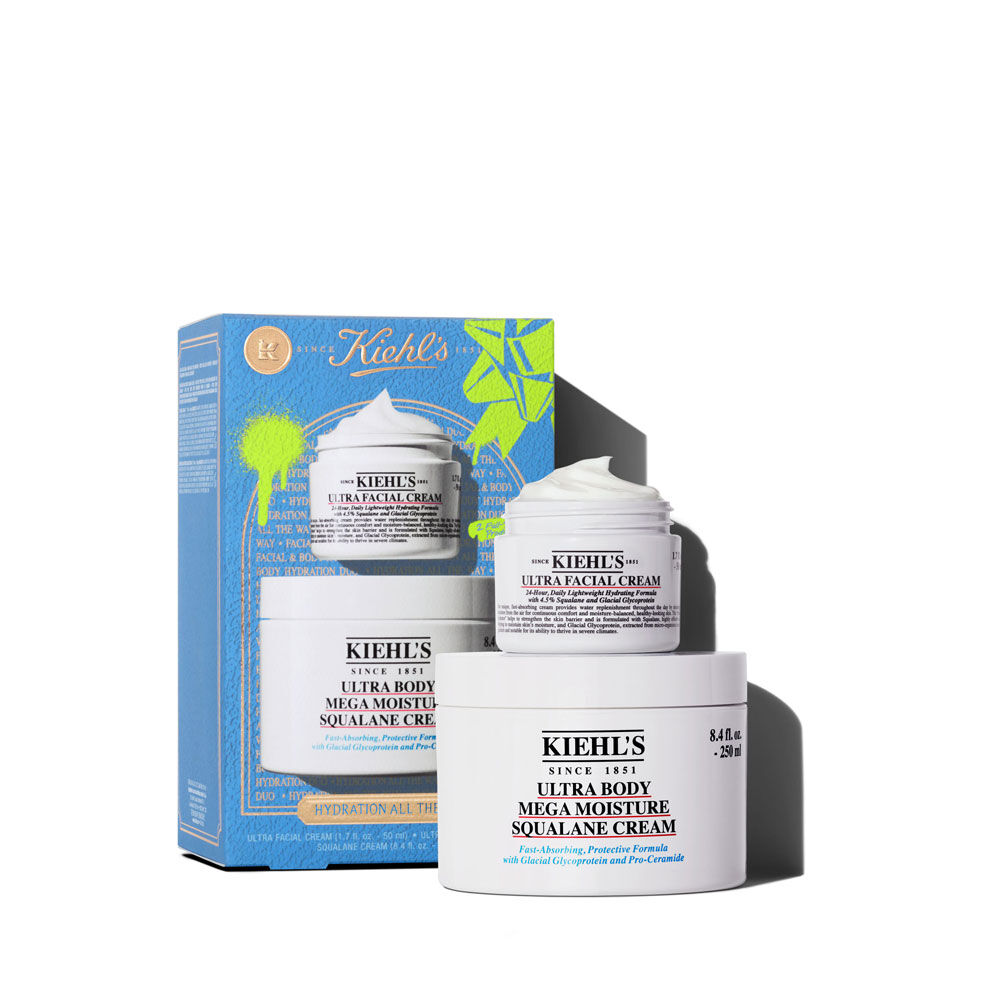 Un grafico muestra una caja de regalo Kiehl’s azul. Contiene dos tarros blancos: "KIEHL’S ULTRA FACIAL CREAM" y "KIEHL’S ULTRA BODY MEGA MOISTURE SQUALANE CREAM". La caja tiene salpicaduras verdes brillantes y un diseno de lazo, con el texto "HIDRATACION COMPLETA" en el frente.