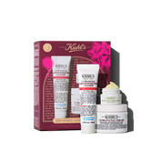 Un grafico muestra una caja de regalo Kiehl's roja a la izquierda. Contiene tres productos: "KIEHL’S ULTRA FACIAL BARRIER-HYDRATING CLEANSER", "KIEHL’S CREAMY EYE TREATMENT WITH AVOCADO" (crema verde palido visible) y "KIEHL’S ULTRA FACIAL CREAM". La caja lleva salpicaduras rosas y un lazo. Texto: "HAVE YOURSELF A HYDRATED HOLIDAY".