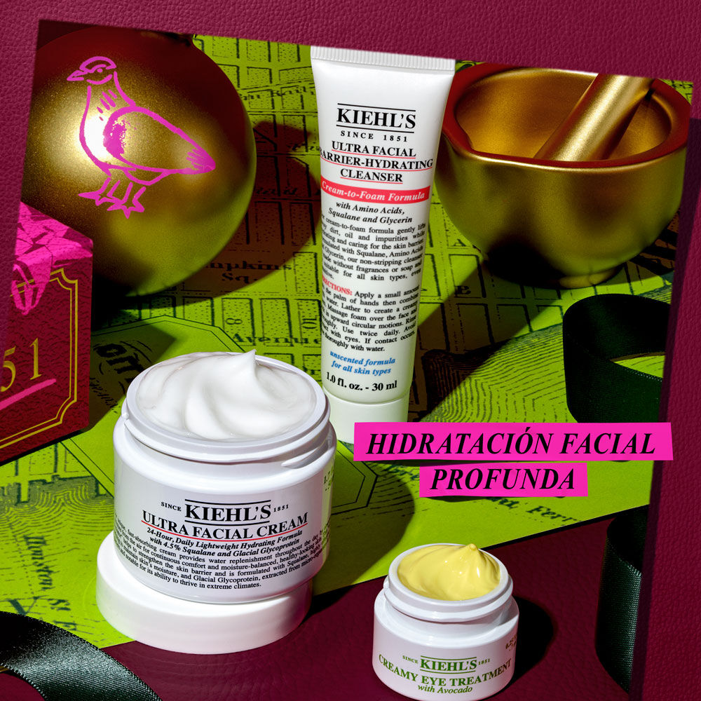 Un grafico festivo sobre fondo rojo oscuro. Muestra varios productos y adornos navidenos: "KIEHL’S ULTRA FACIAL BARRIER-HYDRATING CLEANSER", "KIEHL’S ULTRA FACIAL CREAM" abierto, y "KIEHL’S CREAMY EYE TREATMENT WITH AVOCADO" abierto con crema amarilla palida. Un mortero dorado, un adorno esferico y cinta verde. Una caja rosa dice: "Give the gift of DEEP FACIAL HYDRATION this holiday!".